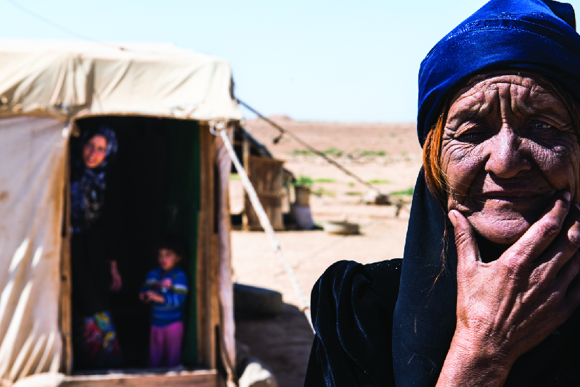 An image of an old person. In the background are an adult and a child in a tent.